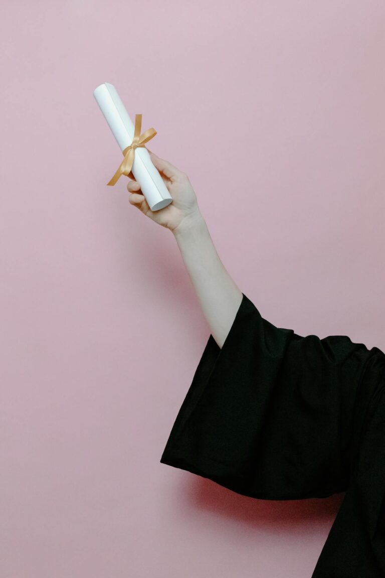 Hand of a graduate wearing a gown holding a diploma with a pink background.